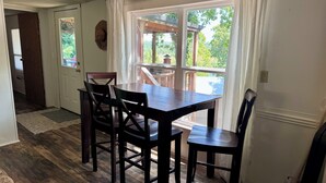 Un coin repas avec une table en bois et quatre chaises, une porte donnant sur une autre pièce, et une vue sur une terrasse en bois et la verdure à l’extérieur.