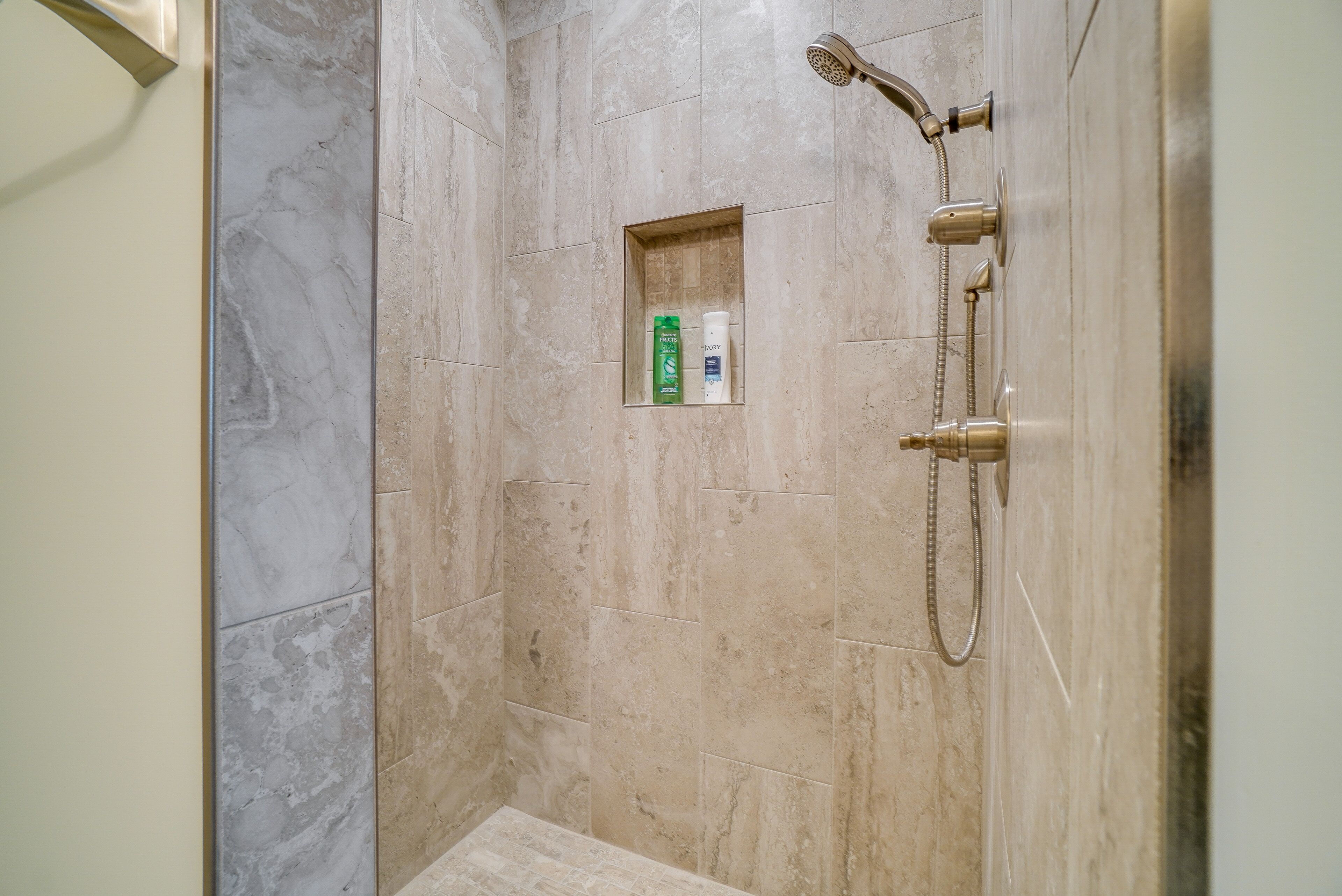 Un área de ducha con azulejos color crema, un cabezal de ducha de mano y un estante empotrado con artículos de aseo.