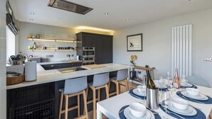 A modern kitchen with a central island, bar stools, and a dining area set for a meal. - Family-Friendly Retreat between Beach & Mountains (Morfa Bychan)