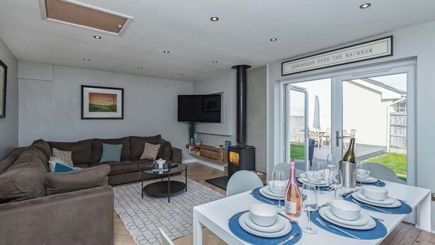 A modern living room with a sofa, TV, fireplace, dining table set for four, and a large window with a view. - Family-Friendly Retreat between Beach & Mountains (Morfa Bychan)