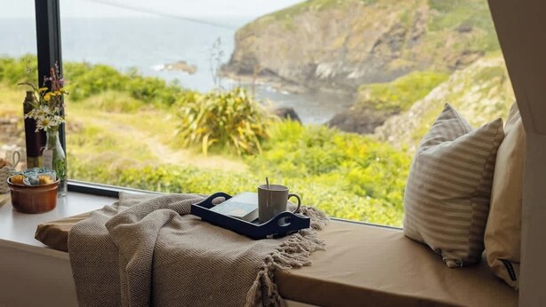 Une vue depuis une fenêtre, avec un plateau contenant une tasse et un livre, un coussin et un vase avec des fleurs.