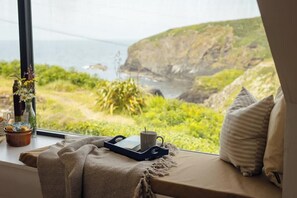 Une vue depuis une fenêtre, avec un plateau contenant une tasse et un livre, un coussin et un vase avec des fleurs.