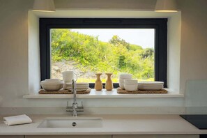 Une cuisine avec une fenêtre donnant sur un espace verdoyant, un évier et une pile d’assiettes blanches posées sur le rebord de la fenêtre.