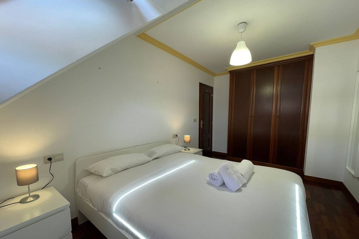 Una cama bien hecha con una toalla enrollada, una lámpara de noche y un armario de madera en una habitación con paredes blancas y suelo de madera.