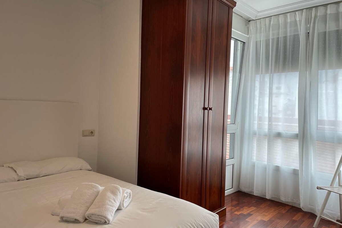 A hotel room with a large bed, a wooden wardrobe, a balcony with sheer curtains, and a wooden floor.