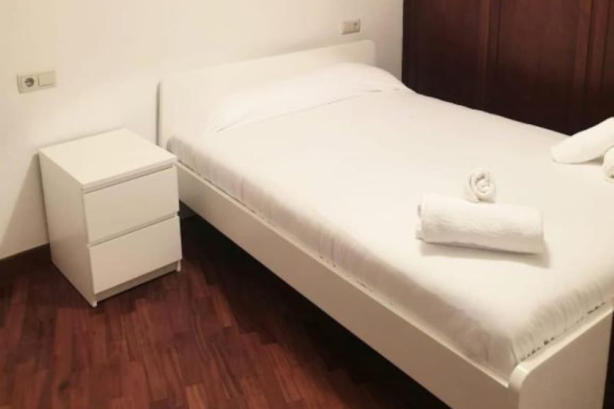 Quarto pequeno e limpo, com uma cama de solteiro, um guarda-roupa de madeira e uma mesa de cabeceira.