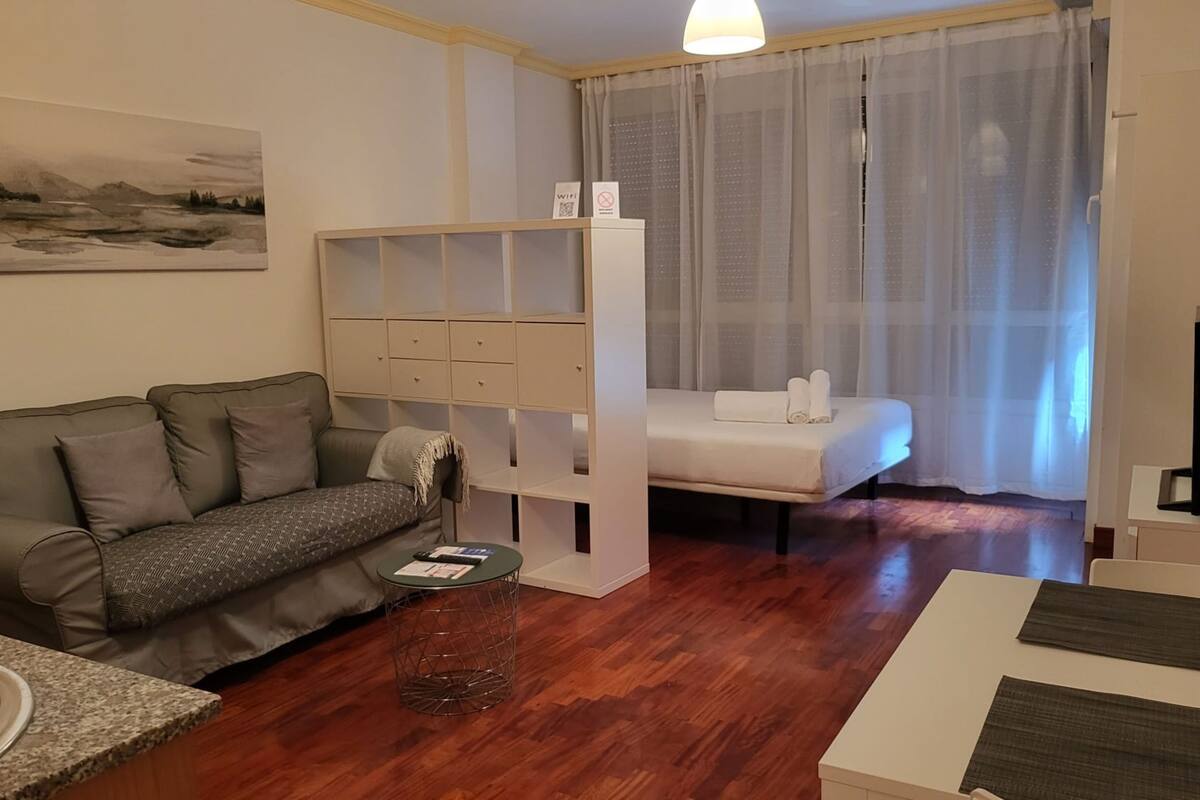 Una sala moderna con un sofá, una mesita redonda pequeña, una cama con cabecero blanco y un ventanal con cortinas de gasa.