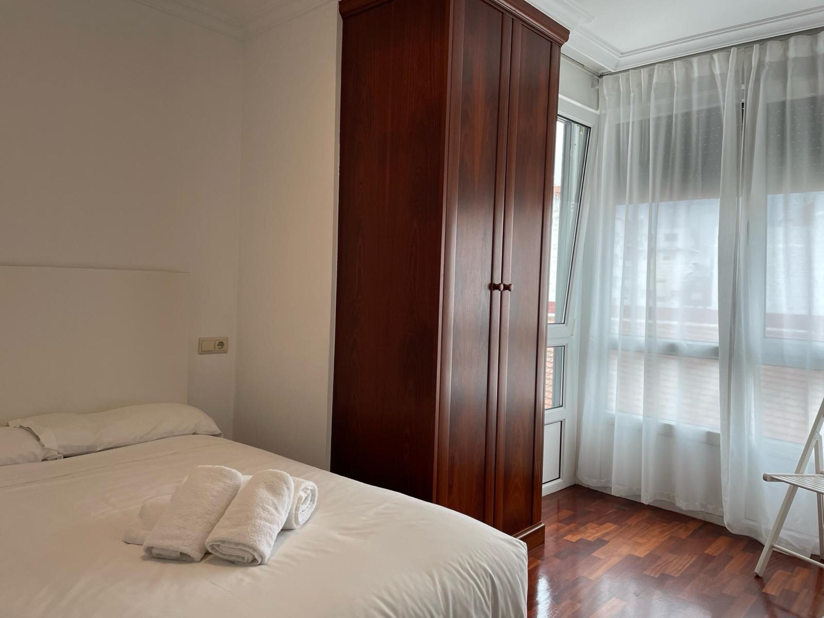 Habitación de hotel con cama, armario, balcón con vistas y ventana con cortinas traslúcidas.