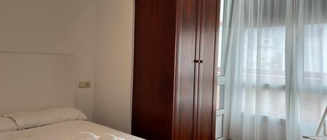 Habitación de hotel con cama, armario, balcón con vistas y ventana con cortinas traslúcidas.