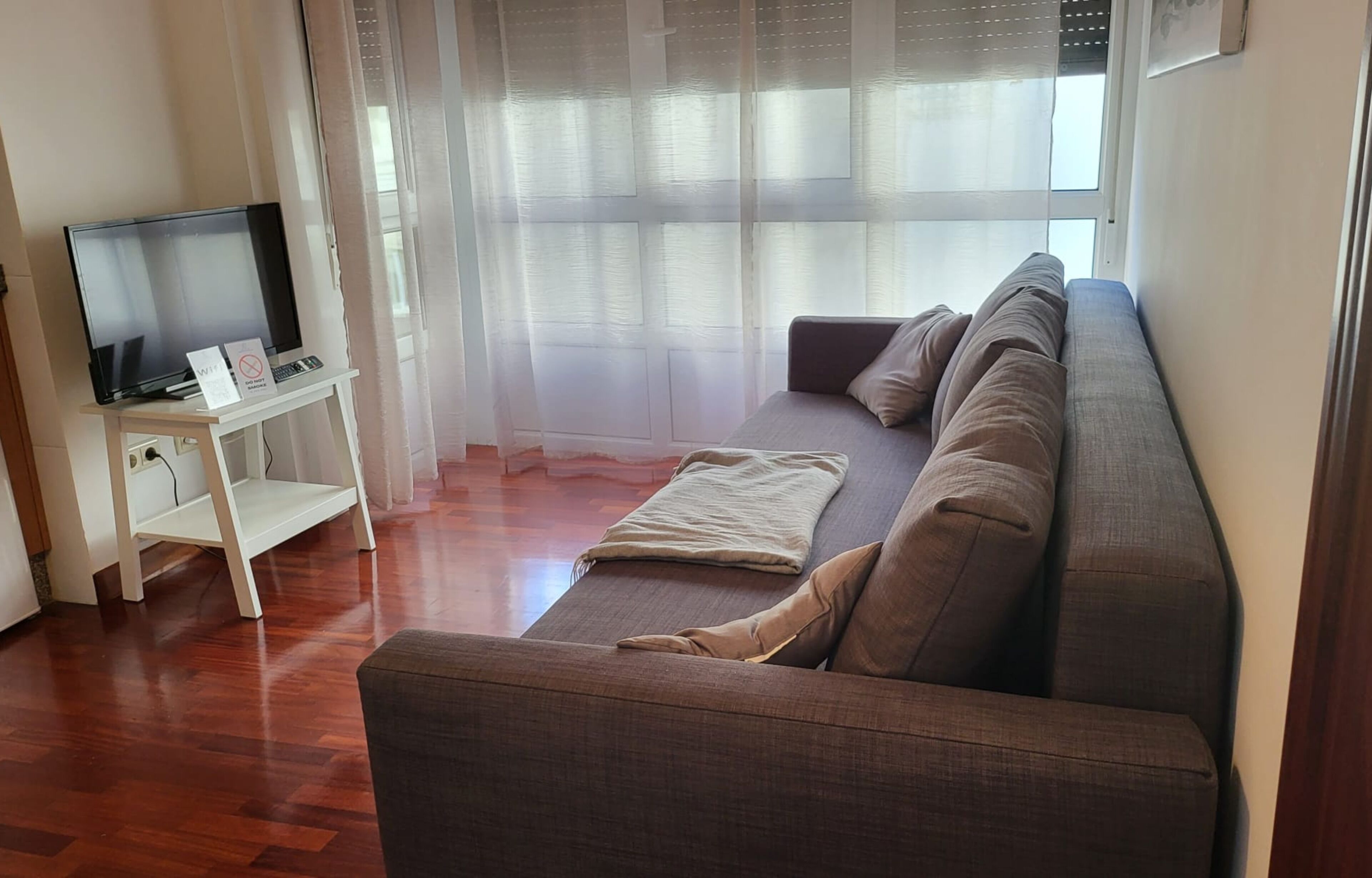 A living room with a sofa, a TV on a stand, and a window with sheer curtains.