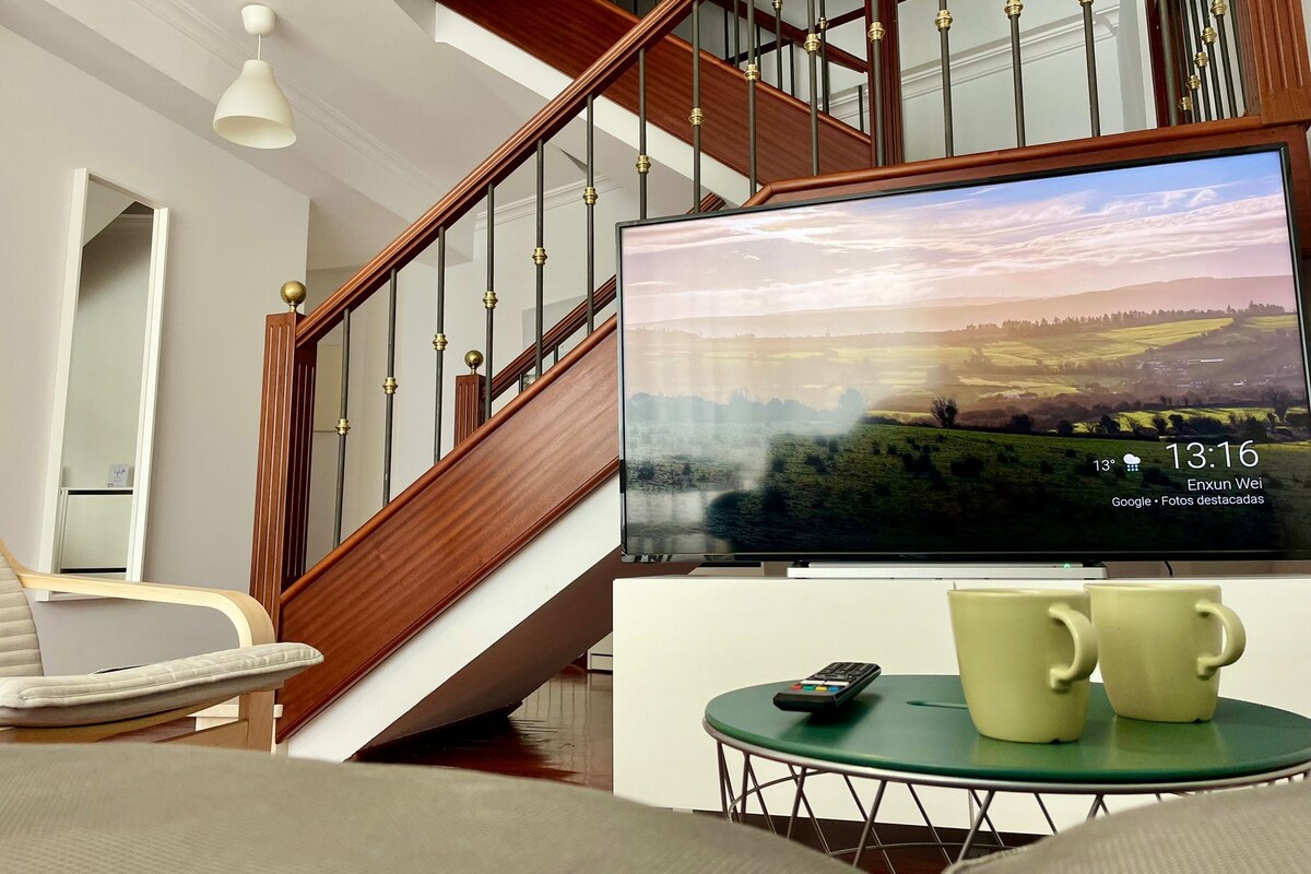 Uma sala de estar com escada de madeira, televisão exibindo uma paisagem e uma mesa verde com duas canecas.