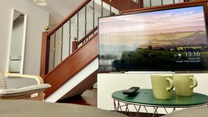 Ein Wohnzimmer mit einer Holztreppe, einem Fernseher der eine Landschaft zeigt und einem kleinen Tisch mit zwei Tassen und einer Fernbedienung.