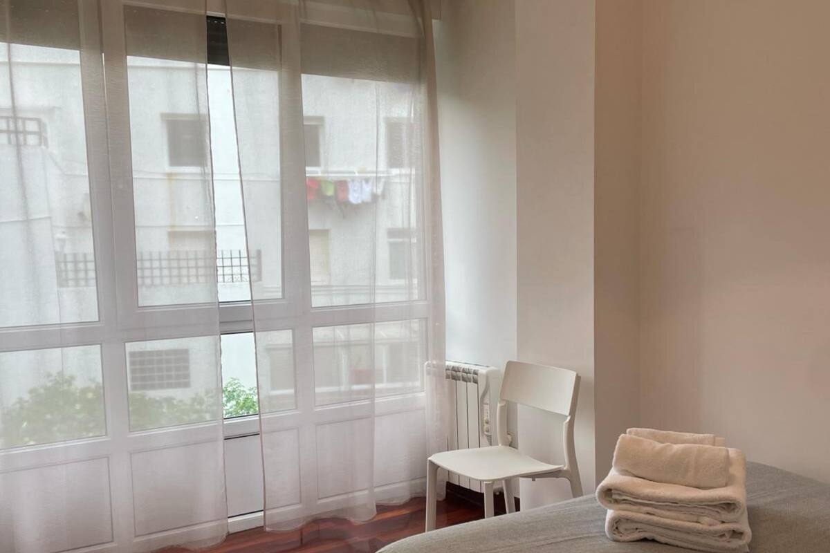 Un dormitorio con cama, una silla, un ventanal con cortinas translúcidas y vista a edificios.