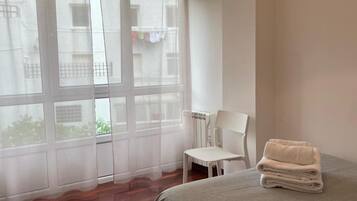 A bedroom with a bed, a chair, a window with sheer curtains, and a view of buildings outside.