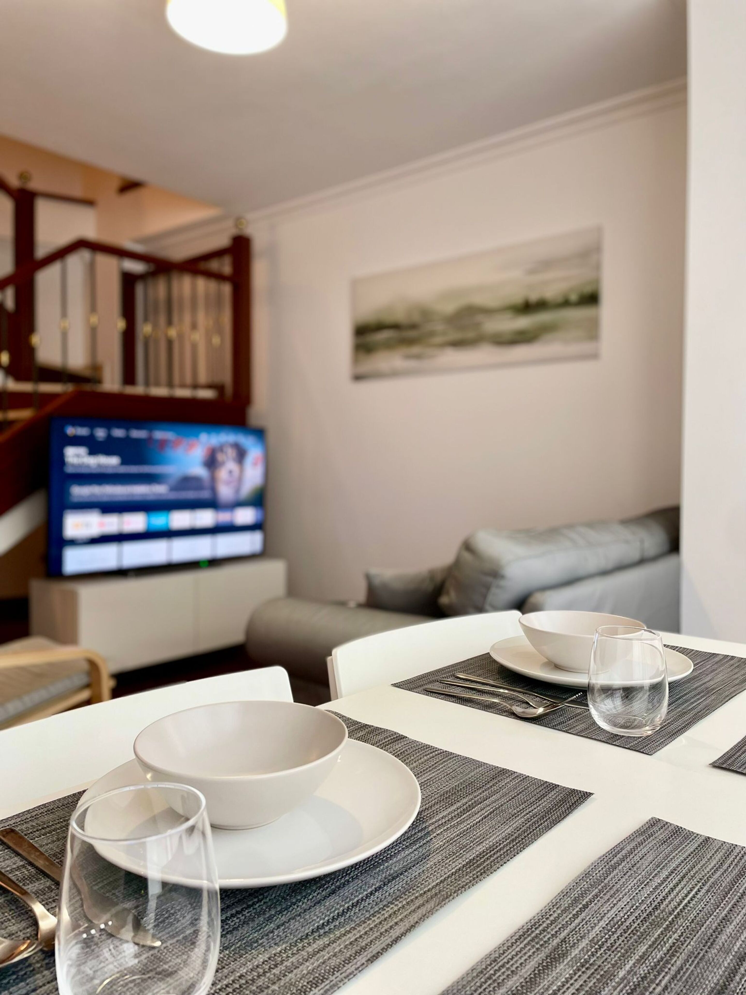 Un coin repas avec une table dressée pour deux personnes, un téléviseur en arrière-plan et un escalier menant à l’étage supérieur.