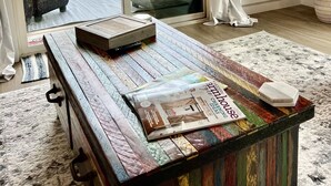 Habitación con un amplio ventanal corredera que da a un espacio exterior con vistas a árboles y un edificio. Hay una mesa de madera con revistas y un control remoto encima.