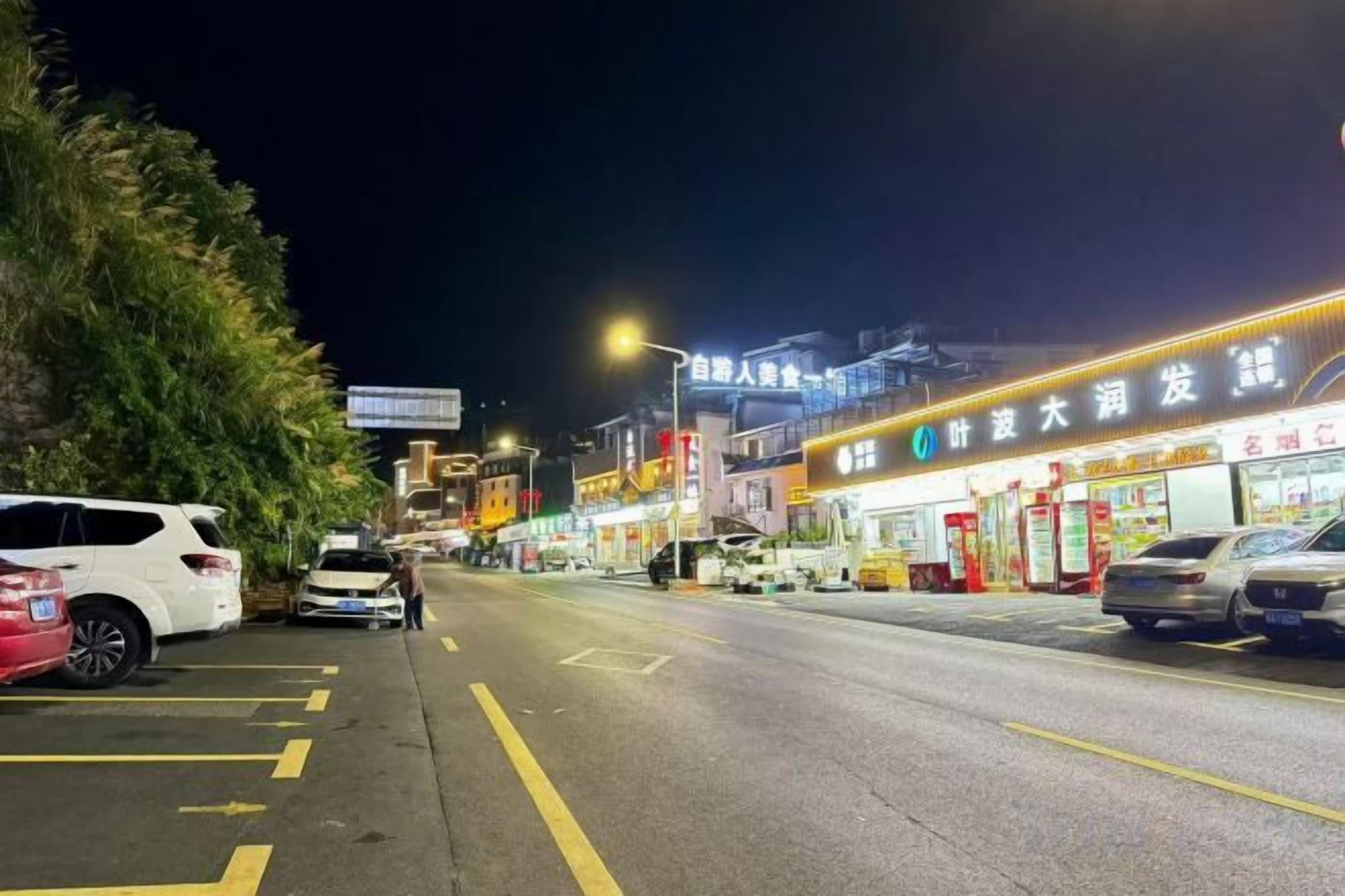 A nighttime street scene with parked cars and illuminated storefronts.