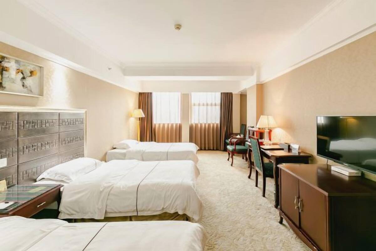 Quarto de hotel com três camas, uma escrivaninha, uma televisão e um armário.