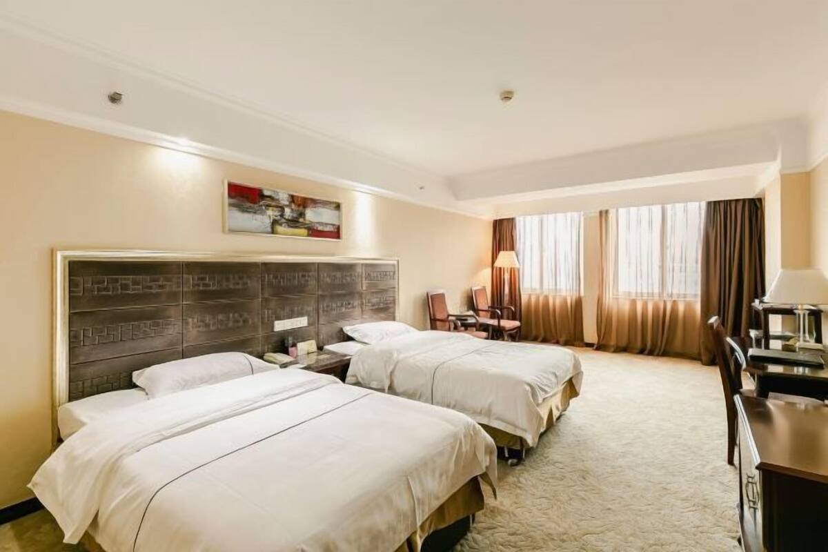 Quarto de hotel com duas camas, uma escrivaninha, uma cadeira, um abajur e um quadro na parede.