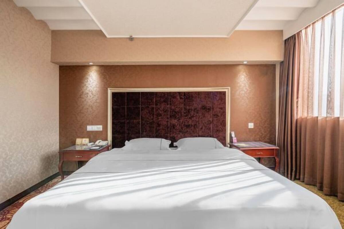 Quarto de hotel com uma cama grande, cabeceira de madeira, mesa de cabeceira, escrivaninha e janela com cortinas.