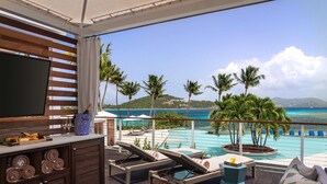A balcony with a TV, a fruit platter, and a bottle of champagne. It overlooks a pool with lounge chairs and palm trees. - Secure Booking Beachfront Ritz 3 Bedroom Property Concierge Services Included (St Thomas)