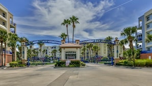 A gated entrance to a hotel complex with a sales office, palm trees, and a roller coaster in the background.