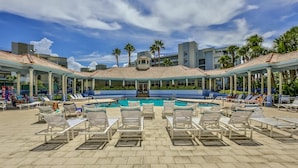 A pool area with lounge chairs and a central pool, surrounded by buildings with a tiled roof and palm trees.
