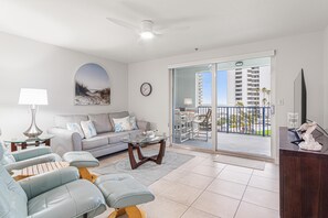 Una sala de estar moderna con sofá, sillones, una mesa de centro, televisor y un balcón con vistas a un edificio de gran altura.