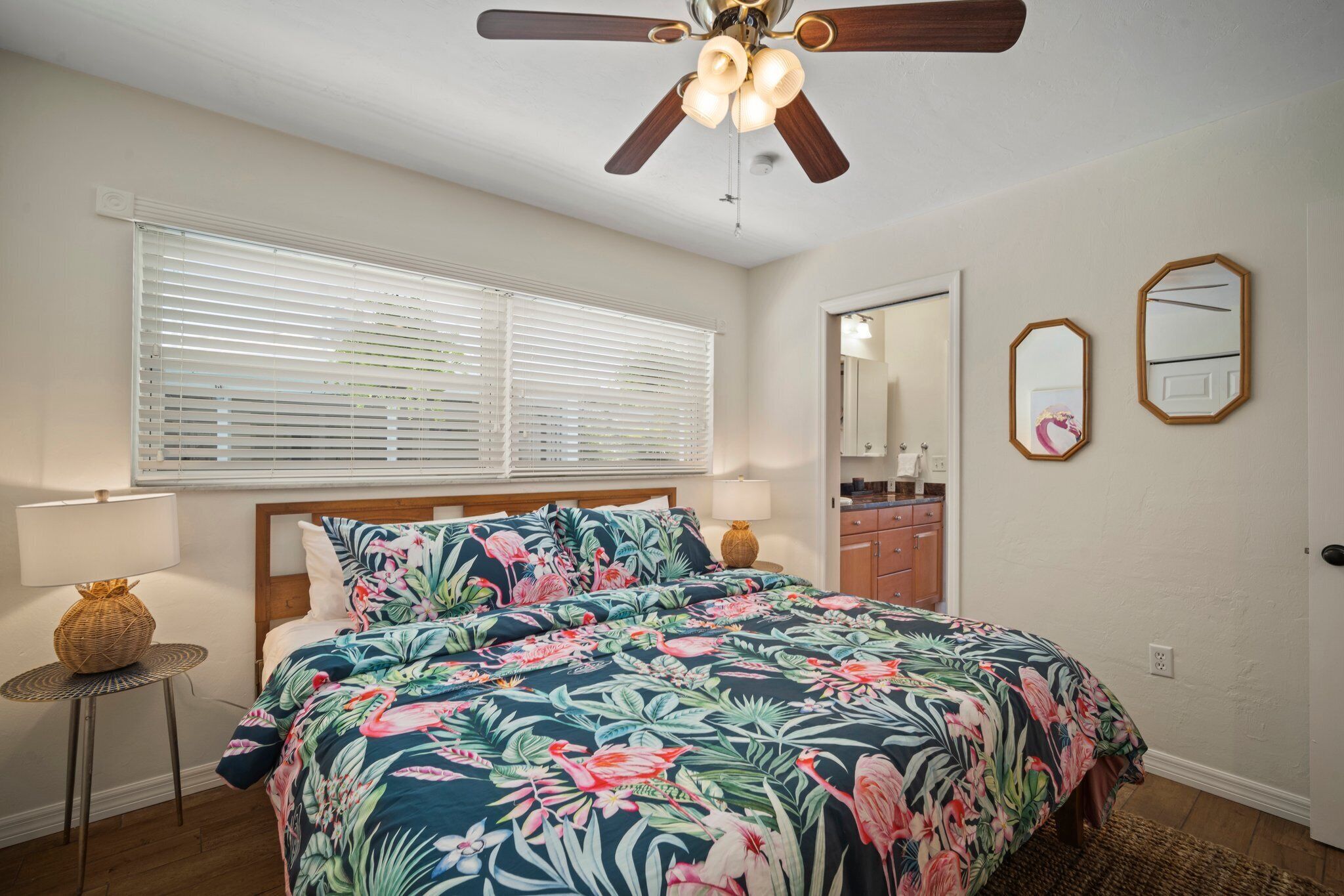 A bedroom with a bed, a ceiling fan, a window with blinds, a bedside lamp, and a mirror on the wall.