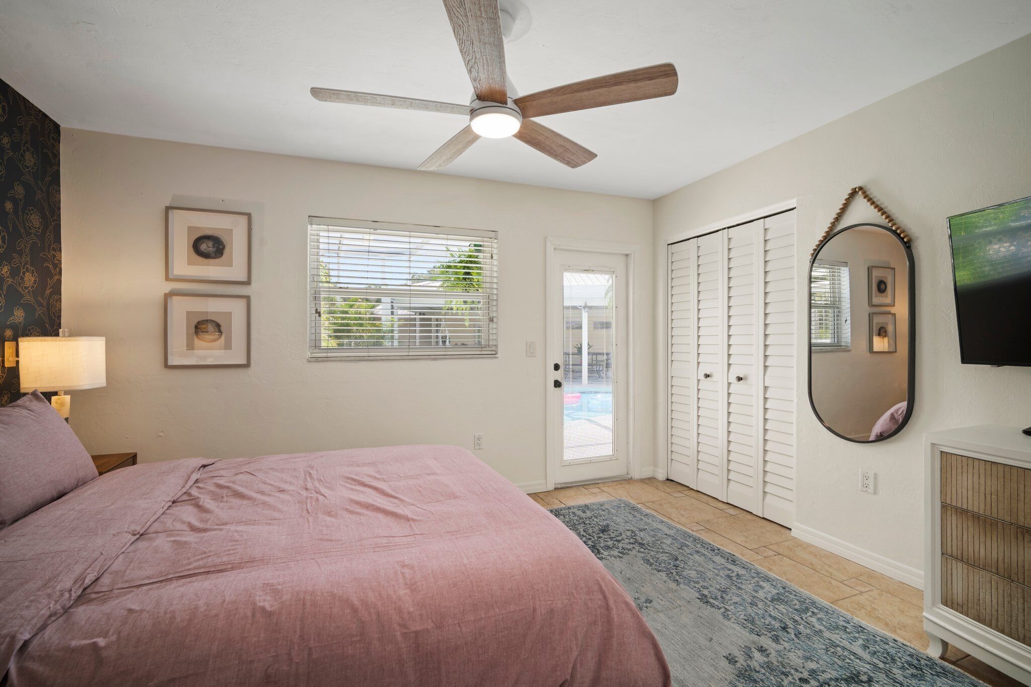 Une chambre à coucher comprenant un lit, un ventilateur de plafond, une fenêtre avec des stores, une porte, un miroir et un téléviseur.