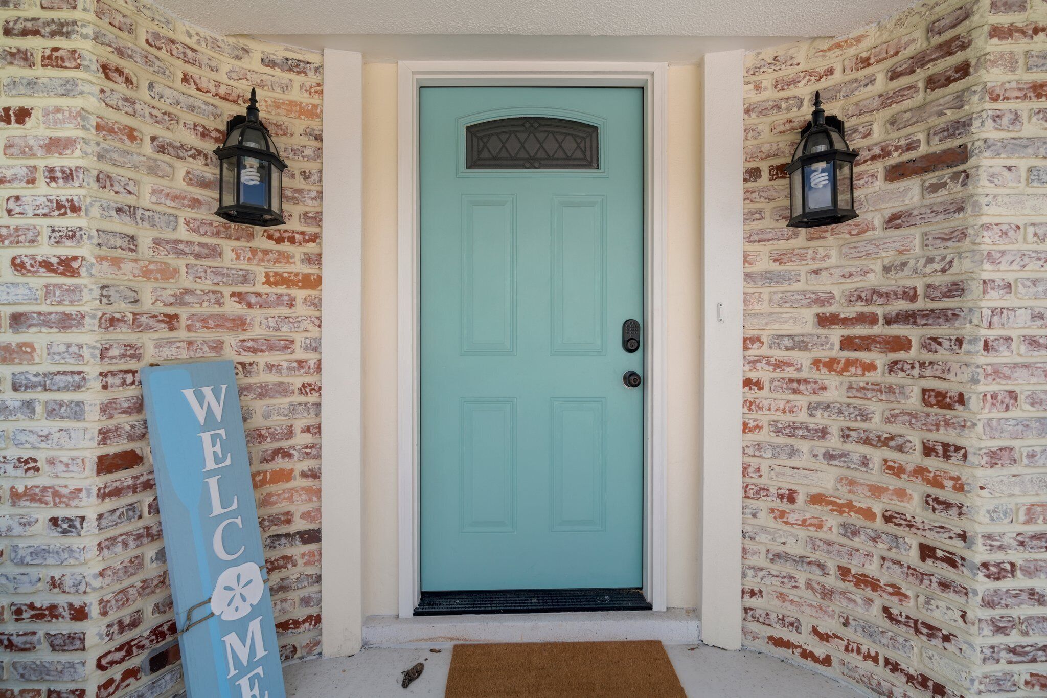 A teal door with a welcome sign, a doormat, and two lanterns on brick walls.