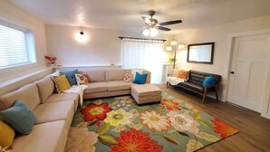 A living room with a beige sectional sofa, a small ottoman, a dark leather couch, a floral area rug, a ceiling fan, and a door. - The White House Hub (Inkom)