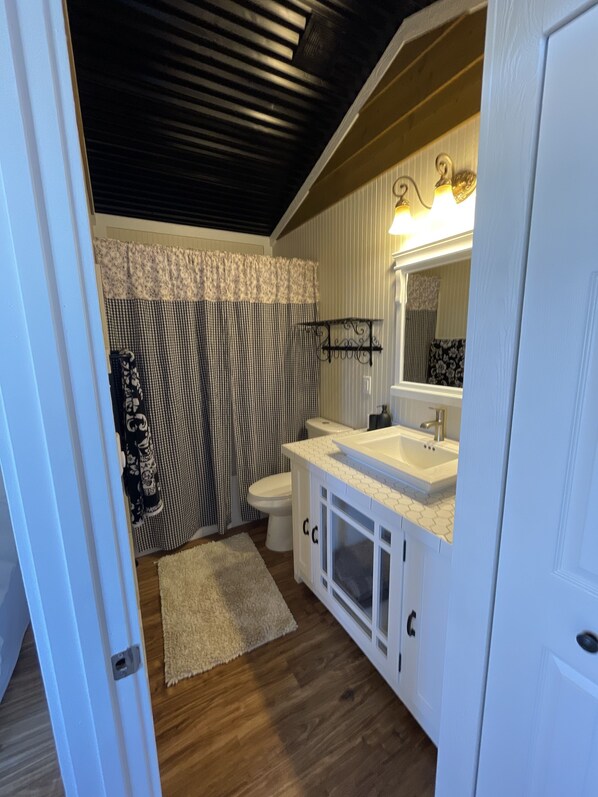 A bathroom with a white sink, toilet, and vanity. There is a patterned shower curtain and a white rug on the floor.