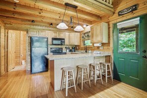 A wooden cabin interior with a kitchen featuring a refrigerator, microwave, and stove, a bar area with stools, and a green door.