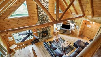 A spacious cabin with a stone fireplace, wooden beams, a ceiling fan, and a cozy seating area.