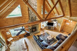 Une cabane spacieuse dotée d’une cheminée en pierre, de poutres apparentes, d’un canapé en cuir et d’un ventilateur de plafond.