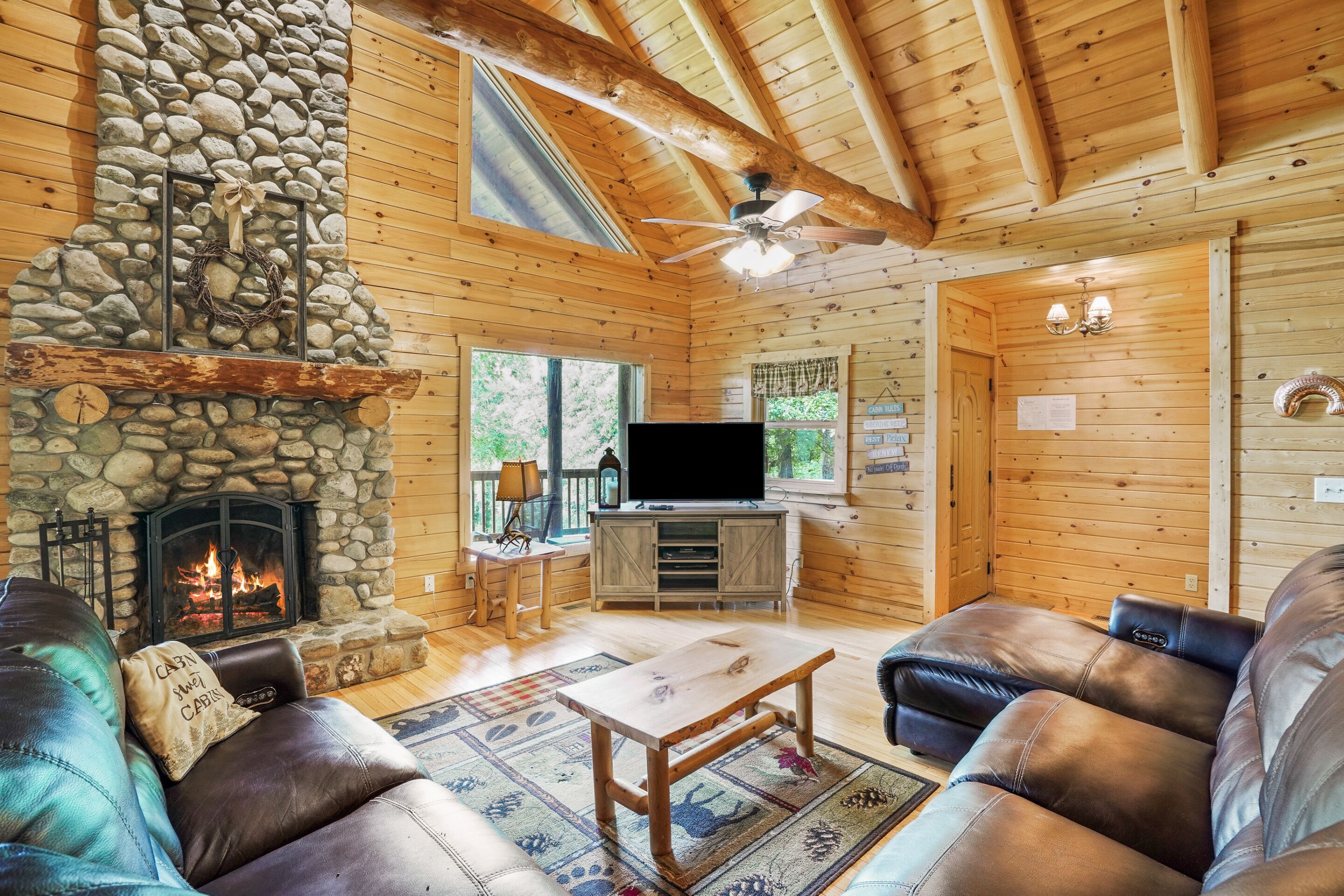 Interior acollidor d'una cabana amb una llar de pedra, sofàs de cuir, un televisor de pantalla plana i un sostre de fusta.