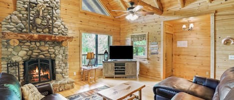 A cozy cabin interior with a stone fireplace, leather sofas, a flat-screen TV, and a wooden ceiling.