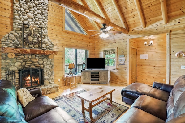 A cozy cabin interior with a stone fireplace, leather sofas, a wooden entertainment center, and a ceiling fan.