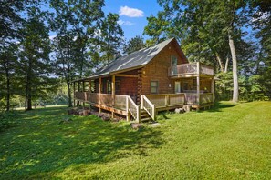 A log cabin with a balcony, surrounded by a grassy area and trees. - Cabin Fever | Hocking, Hot Tub, Pets, Firepit (Logan)