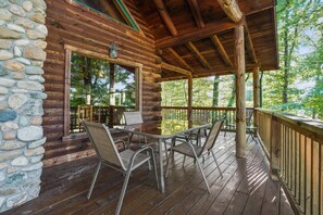 Une cabane en bois avec un mur en pierre, une véranda couverte avec des chaises en métal et une vue sur la forêt.