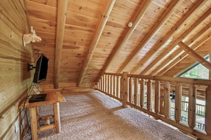 A wooden interior with a sloped ceiling, a wooden desk with a flat-screen TV, and a view through a window.