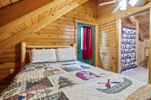 A wooden cabin bedroom with a sloped ceiling, a bed with a quilt, a door with a red curtain, and a wall with a wildlife-themed tapestry.