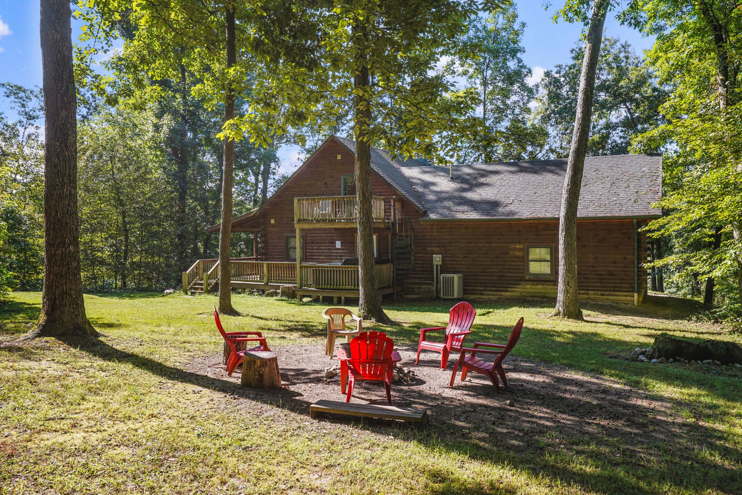 A rustic cabin with a balcony, surrounded by trees and a grassy area with red chairs.