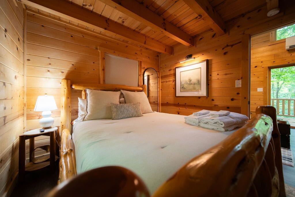 A wooden cabin room with a bed, a nightstand, a framed picture, and a window.