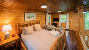 Quarto de cabana de madeira com uma cama grande, mesas de cabeceira, uma televisão e uma janela com cortinas.