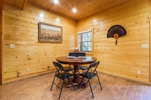 A wooden-paneled room with a round table and four chairs, a framed picture on the wall, and a window with blinds.