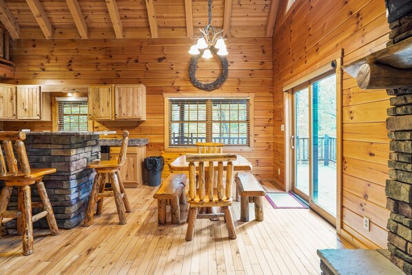 Interior de una cabaña de madera con comedor, chimenea de piedra y ventanas amplias.