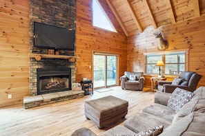 A log cabin interior with a stone fireplace, a television mounted above it, and a leather sofa.