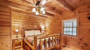 A wooden cabin bedroom with a bed, two bedside lamps, a window with blinds, and a ceiling fan. - Sieri Glen Cabin | Hocking Hills, Sleeps 14 (Logan)